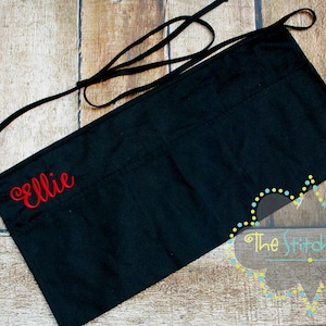 May include: Black apron with a red embroidered name "Ellie" and a white and blue logo that says "The Stitch Shop".