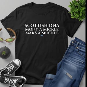 Puede incluir: Camiseta negra con el texto "SCOTTISH DNA MONY A MICKLE MAKS A MUCKLE" en blanco. Se muestra con vaqueros azules y zapatillas deportivas en blanco y negro. También se ven gafas de sol y una pequeña planta.