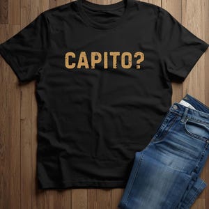 May include: A black short-sleeved t-shirt featuring the word CAPITO? printed in a distressed, tan-coloured font across the chest. The garment is paired with a pair of blue denim jeans, showcasing a classic five-pocket design and a button fly.