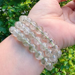 May include: Three clear quartz bead bracelets with green inclusions are displayed. The bracelets are strung on a green elastic cord. The beads are round and have a polished finish. The background is a blurred view of green foliage.