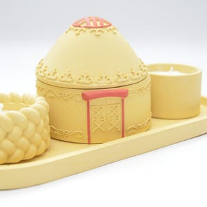 May include: A decorative candle set featuring a yurt-shaped container with a pink roof and door accents, a woven candle holder, and a cylindrical candle, all in a soft yellow hue, presented on a matching tray.