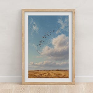 May include: A framed print featuring a landscape scene. The image shows a field of golden wheat under a blue sky with fluffy white clouds. A flock of birds flies overhead. The frame is made of light-colored wood.