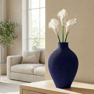 May include: A navy blue vase with a textured, spiral design, holding three white calla lilies. The vase is on a light wood table, with a neutral-toned living room in the background. The vase is approximately 12 inches tall.