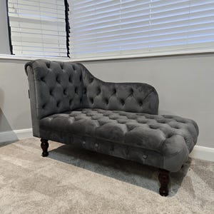 May include: A grey velvet chaise longue with button detailing and dark wooden legs. The elegant furniture has a curved back and armrest. The chaise is on a light grey carpet in front of a window with white blinds.