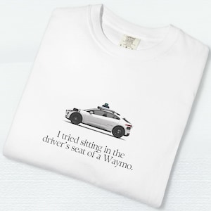 May include: White t-shirt with a graphic of a white Waymo car and the text "I tried sitting in the driver's seat of a Waymo." The shirt has a crew neck and a tag on the collar.