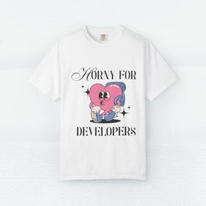 May include: White t-shirt featuring the text "Horny for Developers" above a cartoon heart character wearing headphones. The heart is pink with brown eyes and is holding a phone. The shirt has a crew neck.