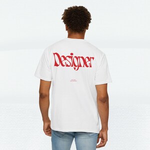 Könnte beinhalten: Weißes T-Shirt mit dem Wort "Designer" in roter, stilisierter Schrift auf dem Rücken. Unter dem Wort "Designer" befindet sich kleinerer Text. Das Shirt ist ein lässiger Stil.