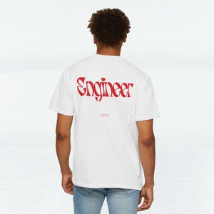 May include: White t-shirt with the word "Engineer" in red, stylized font on the back. Below the word "Engineer" is smaller text. The shirt is a casual, crew neck style.