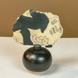 May include: A dark green ceramic jar with a small, round lid. Above the jar is a piece of torn paper with black paint and colorful abstract designs. The jar has a smooth, rounded shape and sits on a white surface.