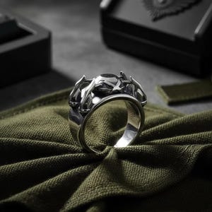 May include: A silver ring with a detailed, ornate design. The ring features a central, dark-colored stone and intricate metalwork around the band. The ring is displayed on a green fabric, with a black box in the background.