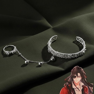 Puede incluir: Un conjunto de cadena de mano plateada que incluye una pulsera y un anillo. La pulsera de brazalete abierto presenta detalles intrincados. Una fina cadena conecta la pulsera con un anillo, también adornado con elementos decorativos. Tres pequeñas esferas colgantes completan el diseño.