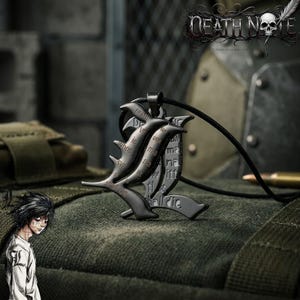 Puede incluir: Un collar con colgante negro con el símbolo "L" de Death Note. El colgante tiene texto grabado y cuelga de un cordón negro. El logotipo de Death Note es visible en el fondo.