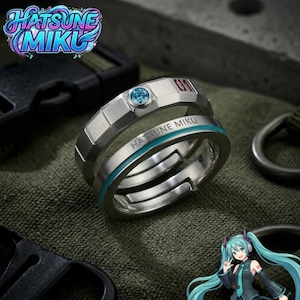 Puede incluir: Un conjunto de anillos de metal plateado que incluye una banda facetada con una pequeña piedra azul y el número 01 grabado en rojo, junto a una segunda banda grabada con el texto HATSUNE MIKU y una fina franja de color verde azulado.