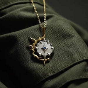 May include: A gold-toned necklace with a pendant featuring a silver-colored, starburst-shaped design with a small blue gemstone at its center. The pendant is accented with gold-colored thorny branches. The necklace is displayed against a dark green fabric.