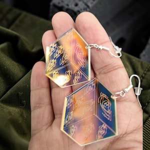 May include: Iridescent geometric earrings with etched designs. The earrings are hexagonal and have a holographic effect, reflecting a spectrum of colors. They hang from silver-colored ear wires, showcasing a unique and modern style.
