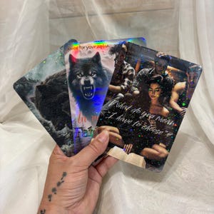 May include: Three holographic cards with fantasy art. One card features a snarling wolf, another has a group of people, and the third shows a wolf in a snowy landscape. Text includes "kne for your ALPHA", "Fat", and "What do you mean I have to choose?"