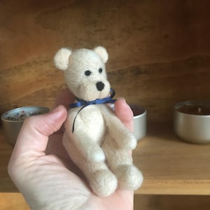 Puede incluir: Un pequeño oso de peluche blanquecino con una cinta azul oscuro atada al cuello. El oso tiene ojos de cuentas negras y una nariz negra. El oso está hecho de un material suave y difuso y se sostiene en una mano.