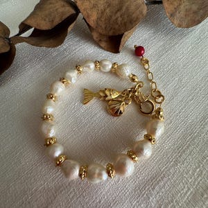 Peut inclure: Un bracelet de perles avec des détails dorés et un charm en forme de poisson. Le bracelet est composé de perles blanches de forme irrégulière avec des perles dorées. Un charm en forme de poisson doré est suspendu au bracelet.