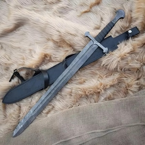 May include: A long, gray sword with a patterned blade and a black handle rests in a black leather sheath. The sword has a cross-shaped guard and a curved handle. The sheath has a strap and buckle. The sword and sheath are on a textured, brown surface.