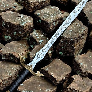May include: A decorative sword with a silver blade etched with a leaf pattern. The hilt has a gold-coloured guard and pommel, and a black wrapped handle. The sword is resting on a bed of brown stones.