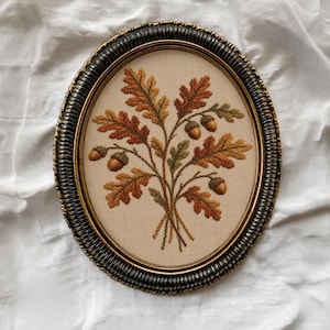 May include: An oval-framed embroidery piece featuring a detailed stitched design of oak leaves and acorns in shades of rust, orange, and olive green. The embroidery is set against a light beige fabric background, enclosed in a dark, textured frame.