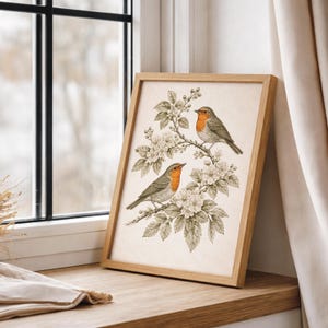 May include: A framed print depicting two robins on a flowering branch. The birds have orange breasts and grey wings. The artwork is set against a cream background, with a light brown wooden frame. The print is displayed on a wooden surface near a window.