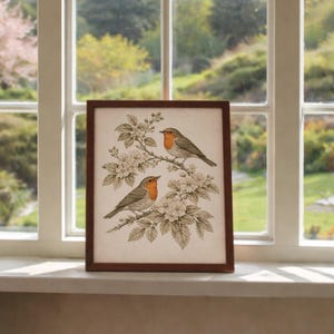 May include: A framed print depicting two robins on a flowering branch. The birds have orange breasts and grey wings. The artwork is set against a cream background, with a dark brown frame. The image is displayed on a white windowsill.