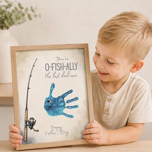 May include: A framed print with a fishing theme. The artwork features a blue handprint fish with a fishing rod illustration. Text reads "You're O-FISH-ALLY the best dad ever" and "Happy Father's Day". The frame is light brown.
