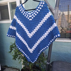 May include: A handmade crochet poncho in blue and white. The poncho features a V-neck design with alternating stripes of blue and white. The garment is displayed on a black hanger, showcasing its open weave and textured pattern. The poncho is approximately 25 inches wide.