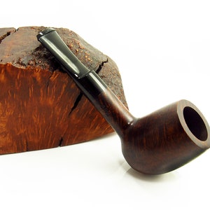 Könnte beinhalten: Eine dunkelbraune Holzpfeife mit schwarzem Schaft, die auf einem Stück Holz mit brauner und orangefarbener Maserung liegt.