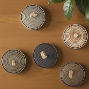 Pode incluir: Cinco bases para copos circulares em vários tons de cinzento, bege e castanho, cada uma com um padrão de ranhuras circulares concêntricas e uma pequena alça de tecido de cor clara fixada ao centro, dispostas de forma dispersa.