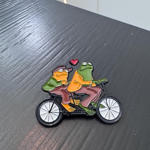 May include: A metal enamel pin featuring two green frogs riding a black tandem bicycle. The front frog wears an orange jacket and brown pants, while the back frog wears a green jacket, brown pants, and a brown cap. A small red heart floats above them.