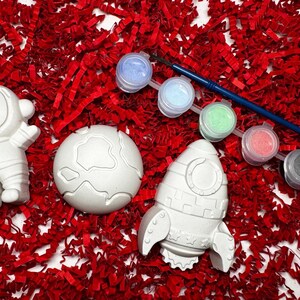 May include: A space-themed craft kit with a white astronaut, planet, and rocket ship figurines. Includes a paintbrush and six paint pots in various colors. The items are arranged on a bed of red paper filler.