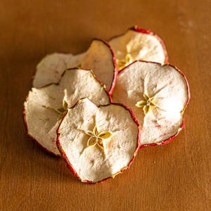 May include: Close-up of several dried apple slices with red edges and a light beige interior. The apple slices are arranged on a wooden surface, showcasing their natural texture and color.