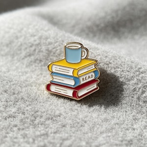 Pode incluir: Um broche de esmalte com borda dourada apresentando uma pilha de três livros com uma caneca azul-clara no topo. O livro superior é amarelo, o do meio é azul com a palavra READ impressa na lateral e o livro inferior é vermelho com páginas brancas.