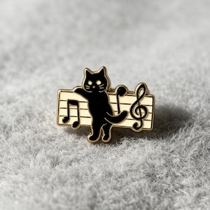 Puede incluir: Un broche de esmalte y metal dorado que presenta un gato negro de pie frente a un pentagrama musical de color crema. El diseño incluye notas musicales negras y una clave de sol, con el gato posicionado centralmente contra las líneas horizontales.