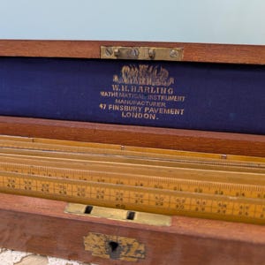 May include: A vintage wooden mathematical instrument set in a wooden box. The box is open, revealing rulers and other measuring tools. The interior is lined with blue fabric. The text "W.H. HARLING MATHEMATICAL INSTRUMENT MANUFACTURER" is visible.