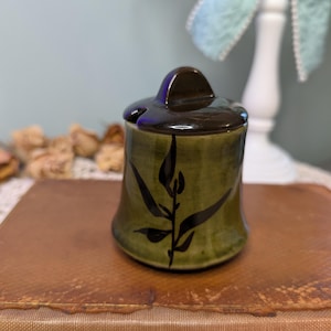 May include: A small, ceramic jar with a lid. The jar is olive green with a dark brown lid and a black leaf design. The jar is sitting on a brown surface.