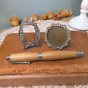 May include: A wooden pen with silver-toned metal accents rests on a textured brown book. Behind the pen are two small, ornate silver-toned metal picture frames, one rectangular and one circular, both featuring detailed floral relief designs.