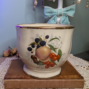 May include: A white ceramic bowl with a gold rim, decorated with a fruit design of peaches, cherries, and blackberries. The bowl is on a brown book, with a lace doily and a lamp in the background.