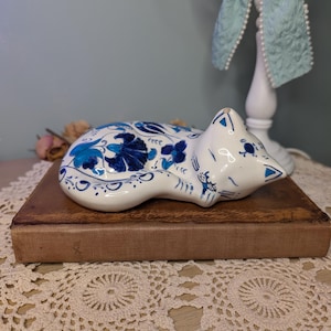 May include: A white ceramic figurine of a sleeping cat decorated with intricate blue floral patterns. The cat is curled in a resting position with its head tucked towards its body, featuring detailed blue painted petals and leaves across its back.