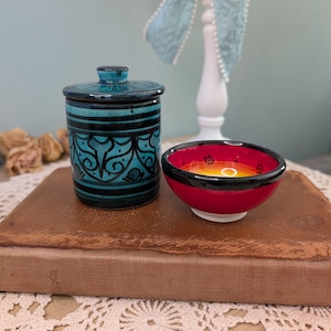 May include: A teal and black ceramic jar with a lid and a small red bowl with a black rim. The jar has a cylindrical shape with a decorative pattern. The bowl has a red exterior and a yellow interior.