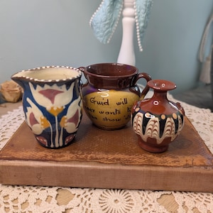 May include: Three small ceramic vases. The vase on the left has a blue, white, and pink floral design. The middle vase is brown with yellow accents and text. The vase on the right is brown with a black and white pattern.