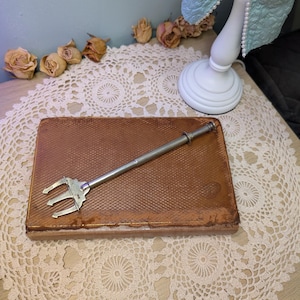 May include: A silver-toned metal trident-shaped serving fork with a long, slender handle resting diagonally across a rectangular, brown, textured vintage book cover. The fork features a three-pronged head and a small, circular ring detail on the handle.