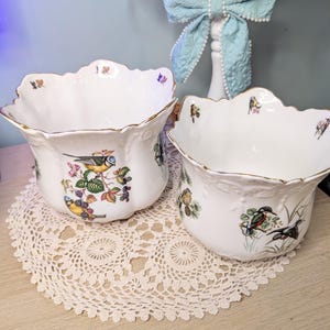 May include: Two white porcelain bowls with scalloped edges and gold trim. Each bowl features a colourful bird and floral design. The bowls are placed on a cream-coloured, crocheted doily.