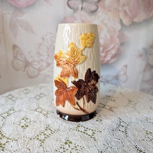 May include: A cylindrical ceramic vase with a cream-coloured, textured bark-like surface. The vase is decorated with raised, three-dimensional maple leaves in shades of yellow, orange, and dark brown. It features a dark brown base rim.