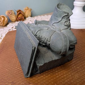 May include: A grey sculpture depicting a pair of boots and an open book. The boots feature intricate lacing and rivets, while the book is open. The sculpture rests on a textured surface, with dried flowers visible.
