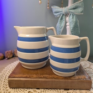 May include: Two ceramic cream jugs featuring a horizontal blue and white striped pattern. Each jug has a curved handle and a pouring spout. The jugs are off-white in colour with bold blue bands encircling the rounded bodies.