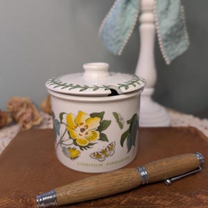May include: A white ceramic jar with a lid, decorated with yellow flowers, green leaves, and a butterfly. A wooden pen with silver accents rests on a brown surface. The jar has the text "GOSSYPIUM BARRADENSE".