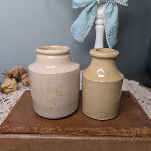 May include: Two vintage stoneware jars in cream and tan shades. The jars have a cylindrical form with a gently widening rim. They are positioned on a brown, textured surface, evoking a vintage or rustic feel. These jars are likely for storage or decorative use.